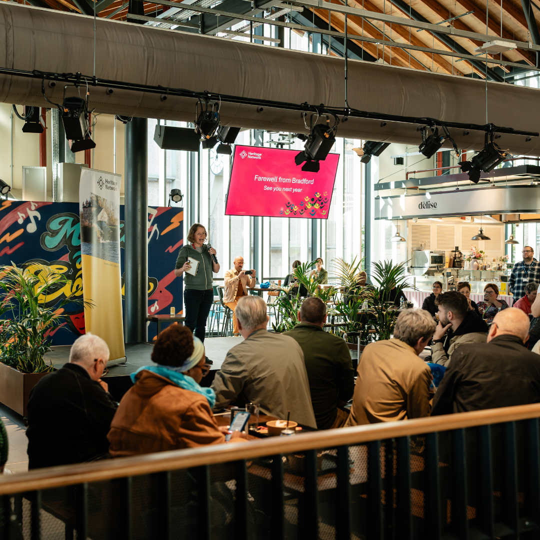 Photograph of groups of people sitting and eating, they are looking at a speaker on a stage.