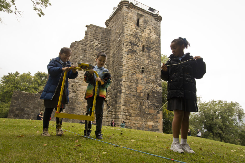 Three schoolchildren use specialist equipment to help survey the grounds around Crookston Castle, the ruins of which can be seen in the background.